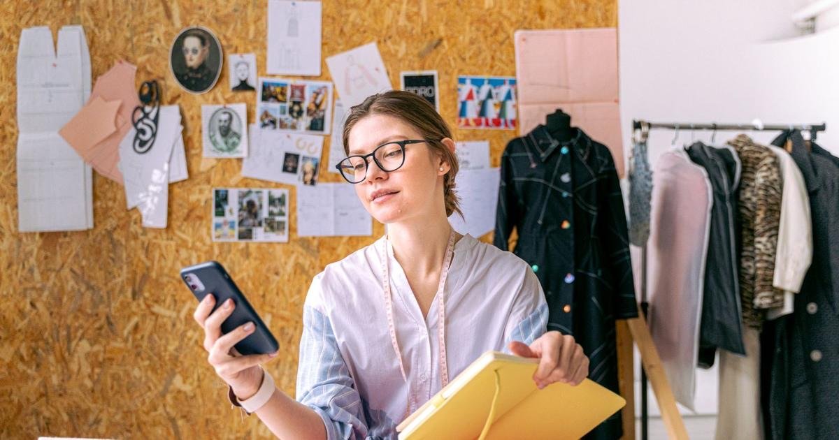 Emprendedora en su taller de ropa revisando su teléfono mientras sostiene una libreta
